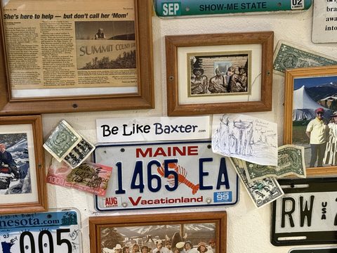 A Be Like Baxter bumper sticker pinned to a wall crowded with vintage licence plates, framed photographs, signed dollar bills and newspaper clippings