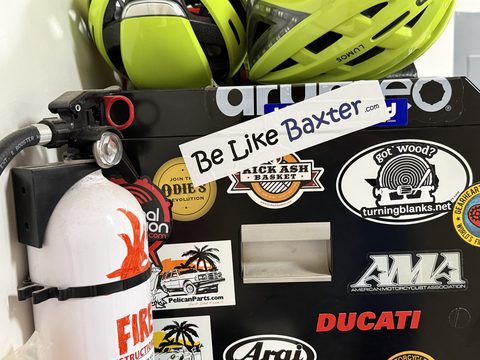 A Be Like Baxter bumper sticker among motorcycle stickers on a workshop toolbox, beside a cycling helmet and a fire extinguisher